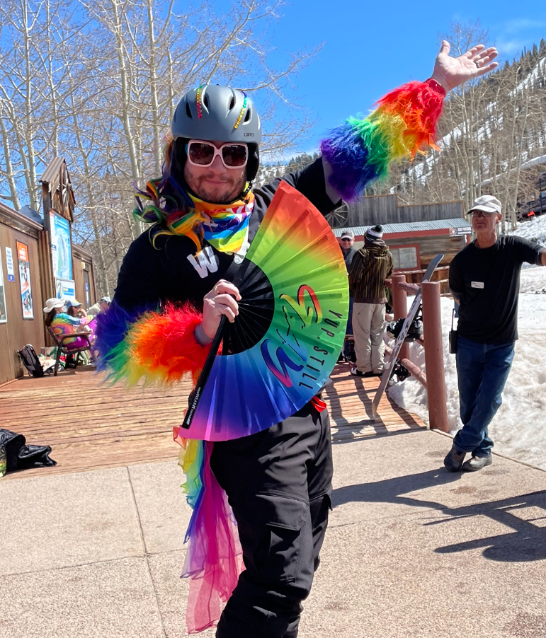 Minister LaLa at a ski resort holding a rainbow fan, rainbow fuzzy sleeves, and a rainbow tutu.